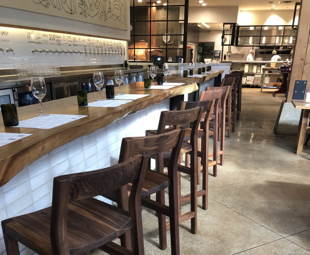 A bar seating area with handcrafted wooden stools and a live-edge wood counter designed by South Loft, in a modern restaurant setting.