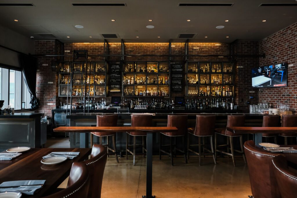 South Loft custom walnut bar tables with leather barstools, facing an elegant brick-lined backlit bar with a variety of spirits on display.