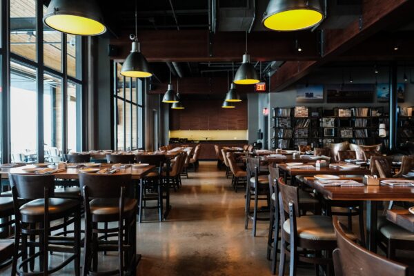 A modern dining room furnished by South Loft with wooden tables and leather chairs, illuminated by large industrial pendant lights. The space features floor-to-ceiling windows on one side, letting in natural light.