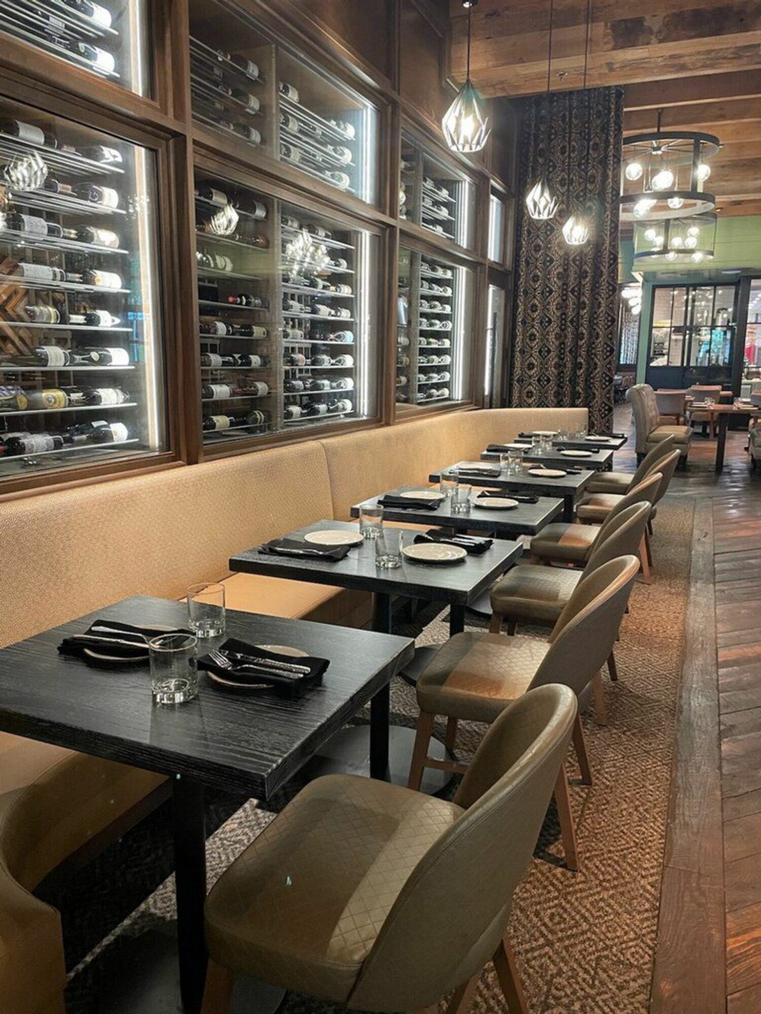 A sophisticated dining area furnished by South Loft featuring a row of modern black tables paired with elegant upholstered chairs. The backdrop includes a well-lit wine display encased in glass, showcasing an extensive wine collection, complemented by warm lighting and luxurious decor.