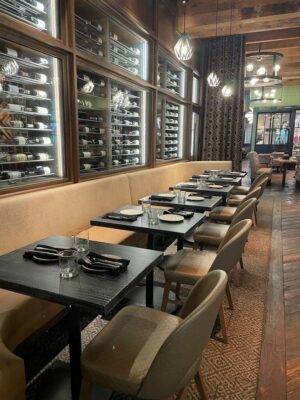 A sophisticated dining area furnished by South Loft featuring a row of modern black tables paired with elegant upholstered chairs. The backdrop includes a well-lit wine display encased in glass, showcasing an extensive wine collection, complemented by warm lighting and luxurious decor.