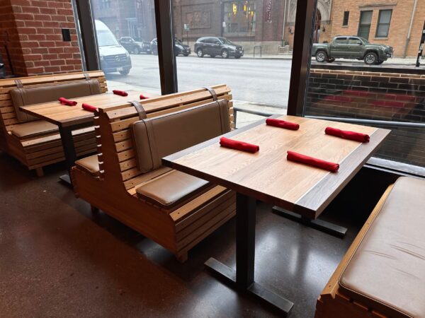 South Loft custom wooden booth seating with light brown cushions, paired with a matching wooden table featuring four red napkins, in front of a large glass window with a street view.