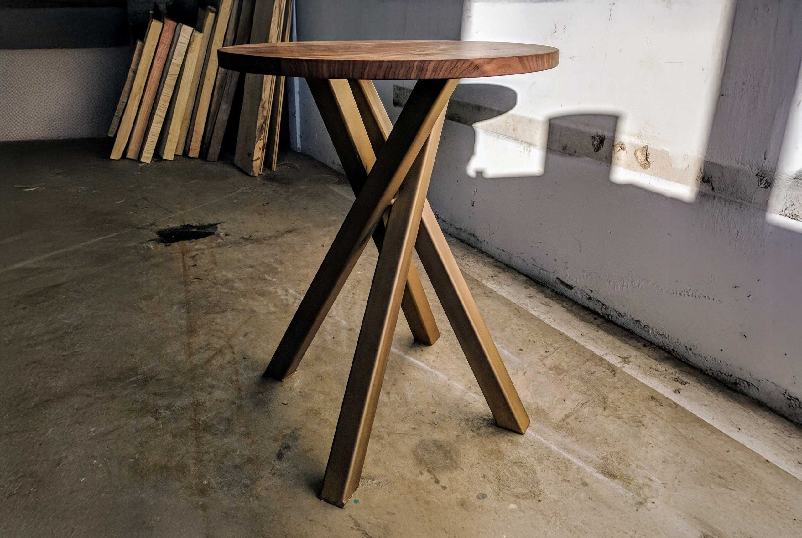 A hardwood side table created by SouthLoft sits in the production facility. It features a round top and a scattered-sticks style base.