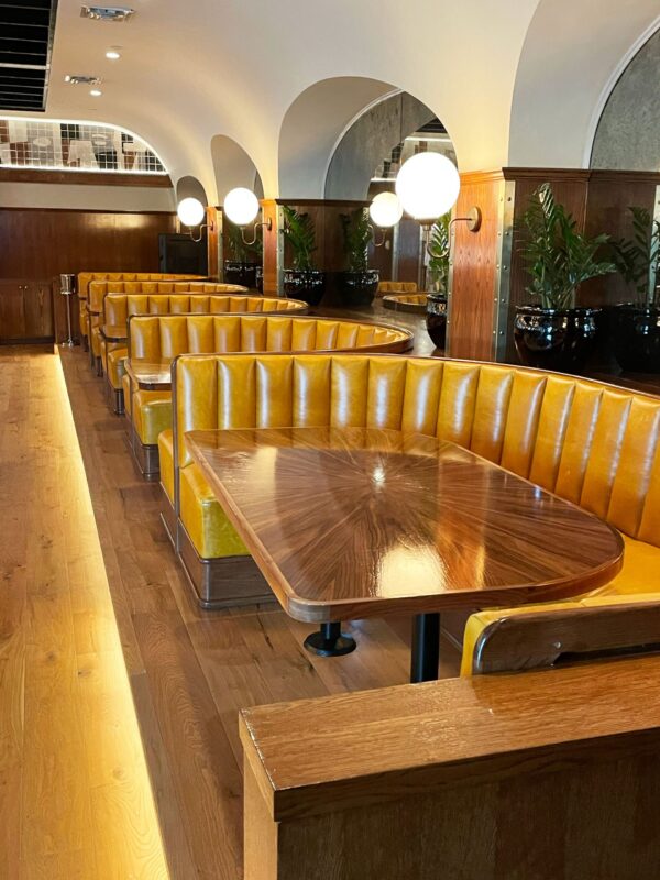 A row of booth style, starburst walnut tables with metallic banding and fasteners sits in Goodwins Dallas. The yellow leather compliments the warm wood tones.